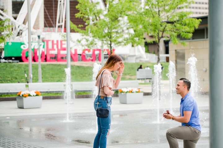 young man in blue polo on one knee proposing to young woman's finger in front of Ferris wheel in cincinnati ohio