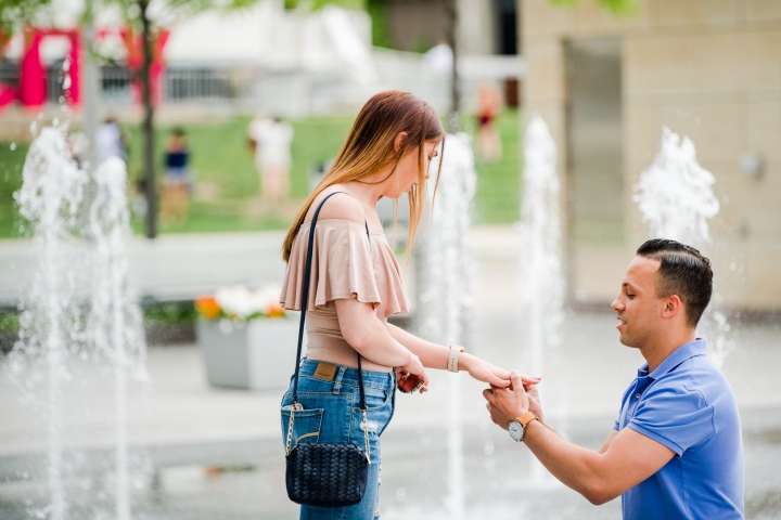 young man in blue polo placing engagement ring on young woman's finger in front of Ferris wheel in cincinnati ohio