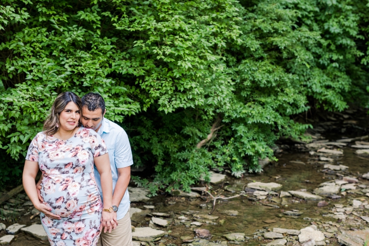 Hispanic pregnant woman  in a blue floral dress standing in creek bed with her husband who is kissing her shoulder while holding hands