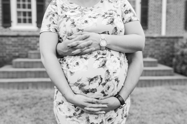 black and white photo of a Hispanic man and pregnant woman  in a blue floral dress while intertwining hands together around her belly.