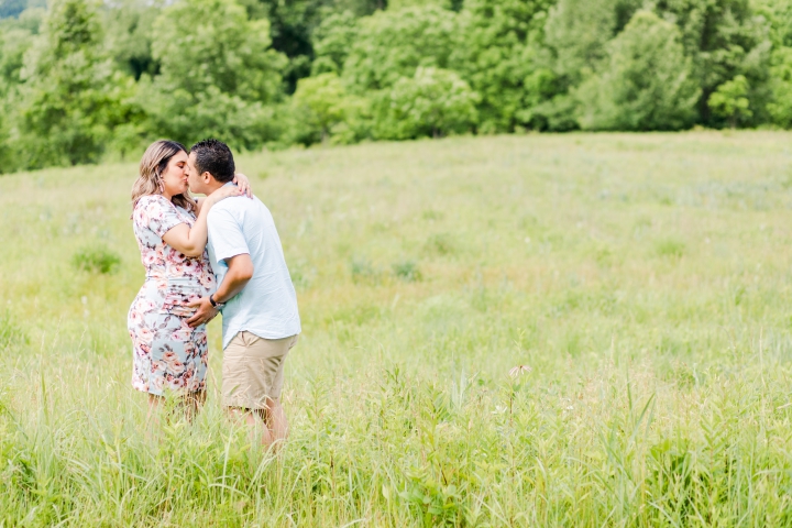 Hispanic man and pregnant woman  in a blue floral dress kissing in the field as he holds onto her belly