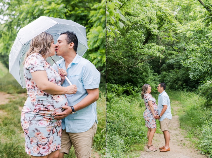 Hispanic man and pregnant woman  in a blue floral dress holding clear umbrella kissing and laughing on park trail