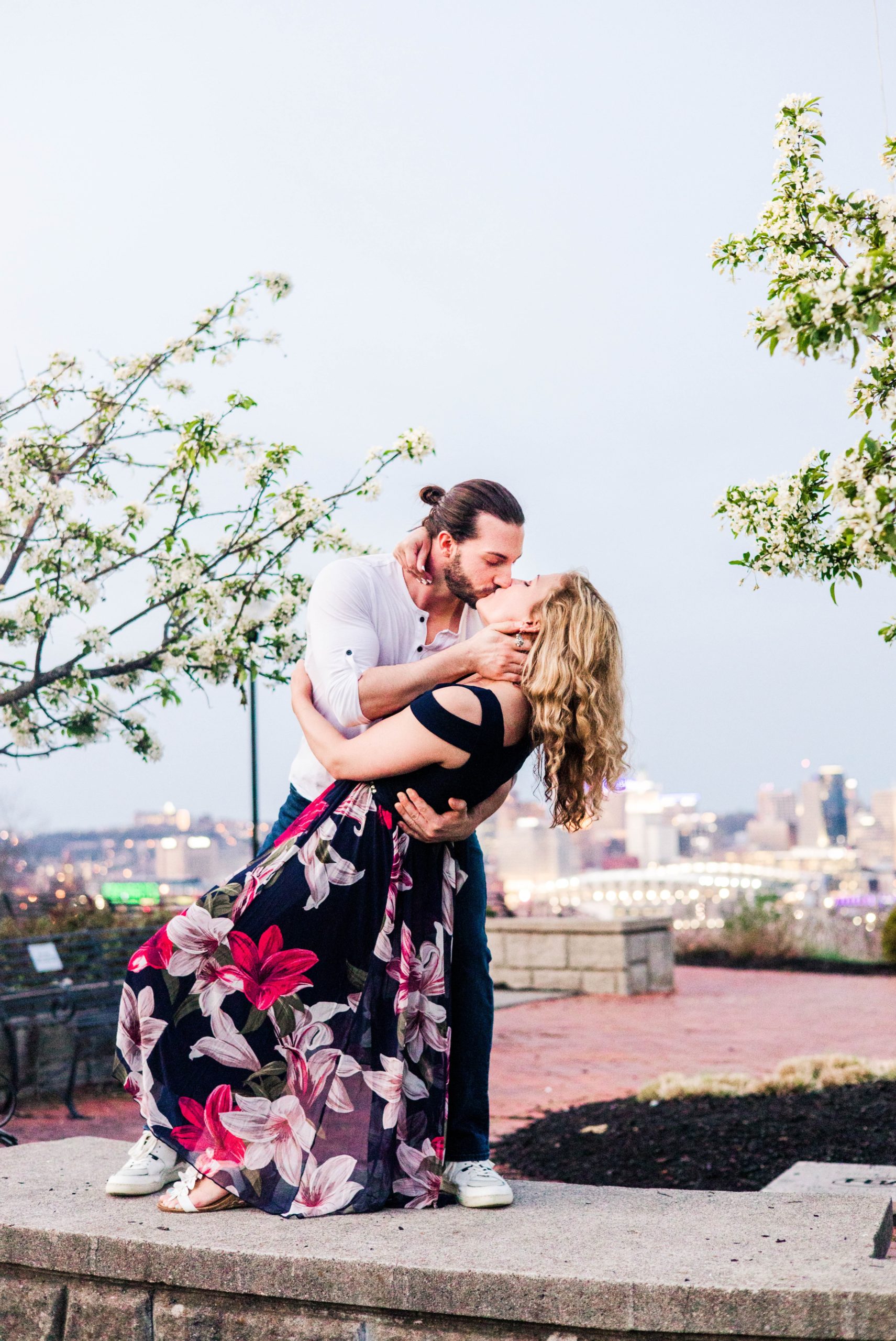Stacy and Joseph standing on a low wall and kissing with the city in the background during their spring engagement session