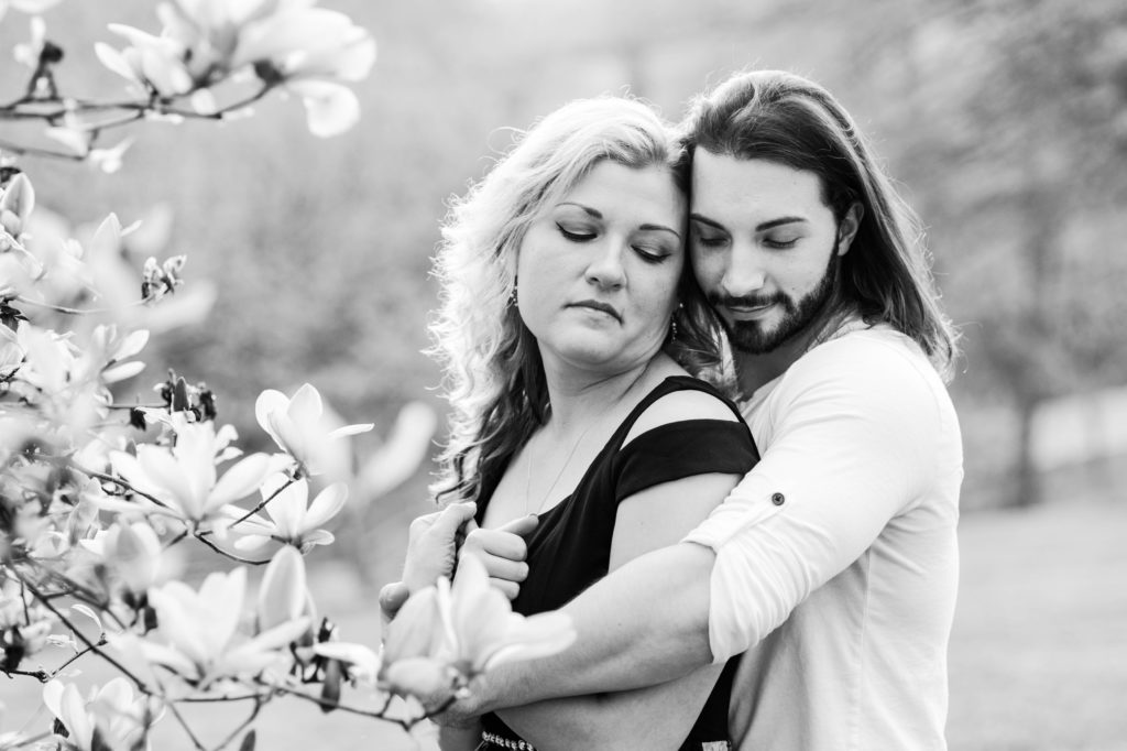 Black and white picture of Joseph hugging Stacy from behind with flowers in the foreground during their The couple kissing in front of some greenery during their spring engagement session