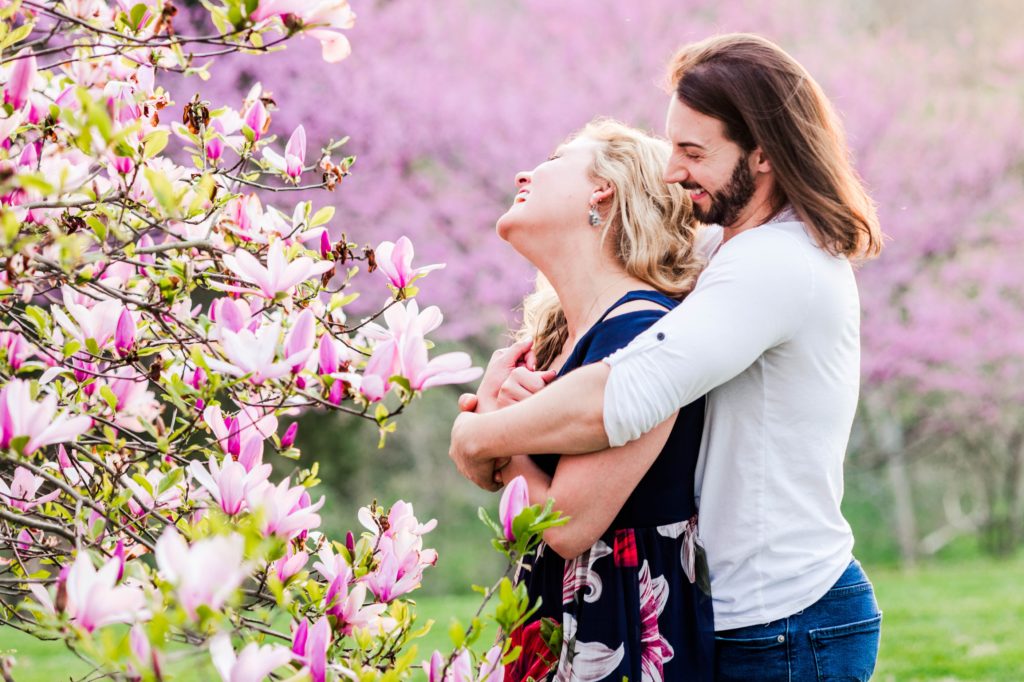 Joseph hugging Stacy from behind while she laughs in front of a tree filled with flowers during their The couple kissing in front of some greenery during their spring engagement session