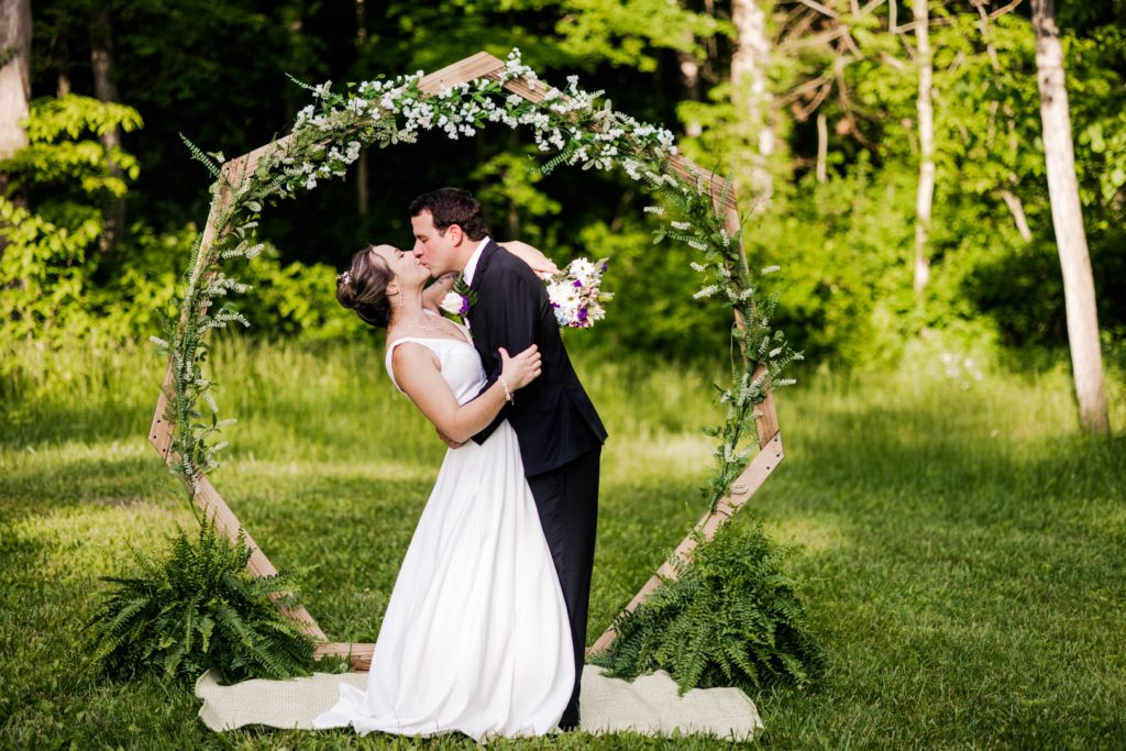 Abby and Ross sharing a kiss in front of a floral arch in a wooded opening at Hueston Woods for their spring wedding.