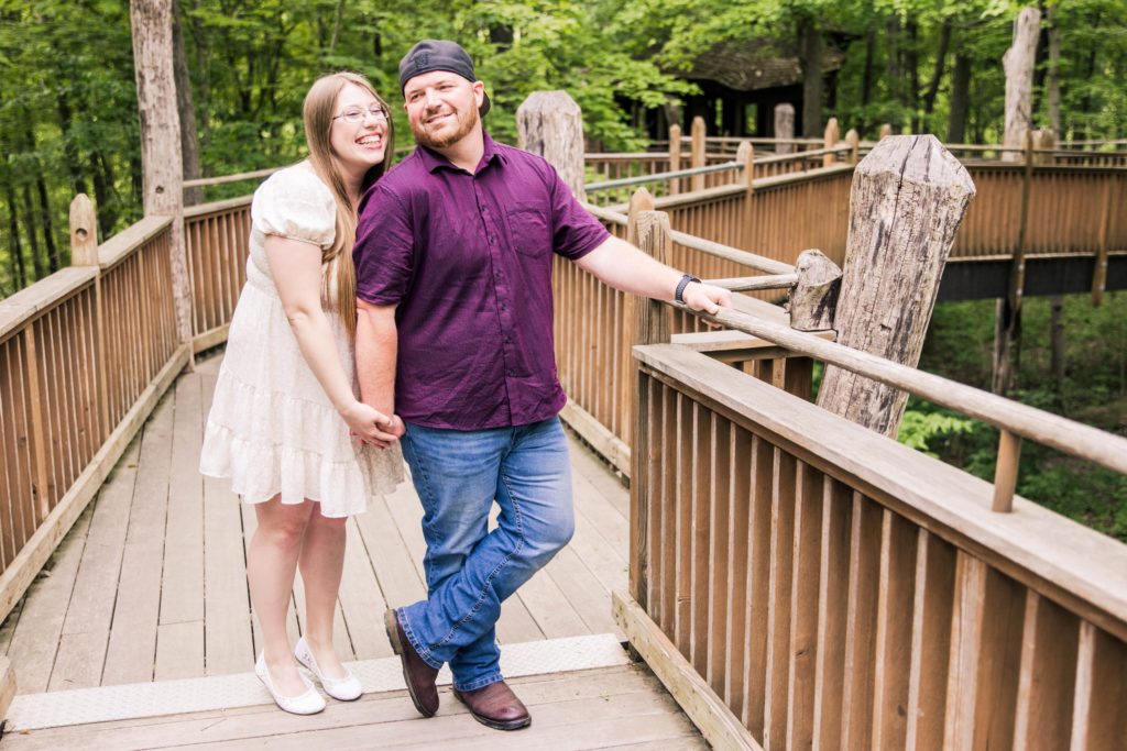 On the walkway of Mt Airy Forest Treehouse, the engaged couple are holding hands as the bride to be laughs at her groom to be who is smiling back at her.