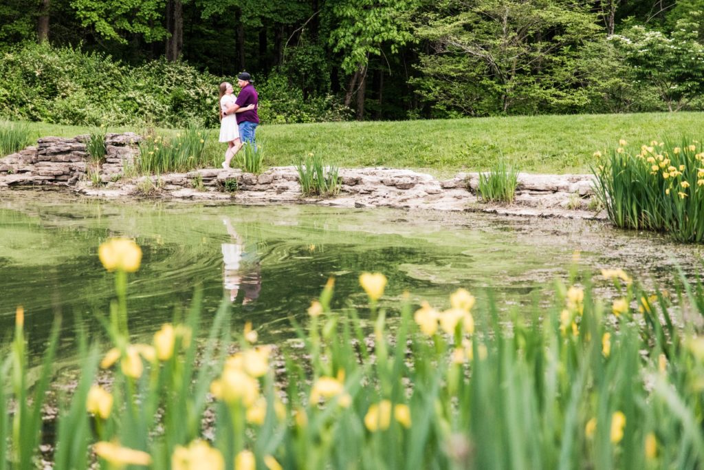 Wide photo of the pond at Mt. Airy Forest with Jasmine and Brandon standing facing each other holding around the waist 