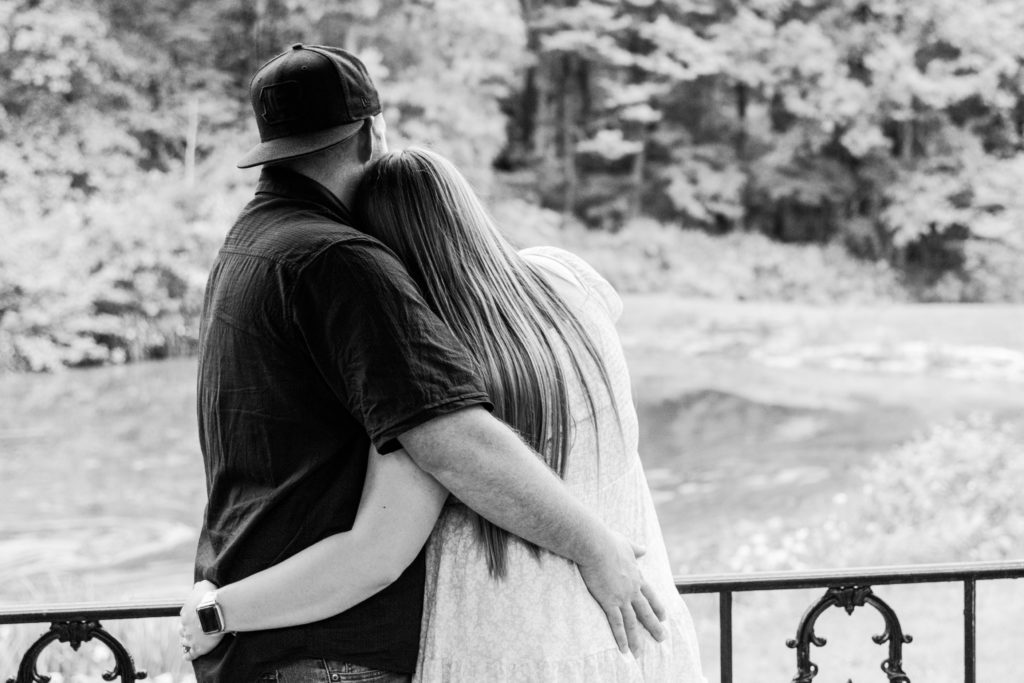 Black and white photo of couple hugging from behind in the gazebo at Mt. Airy Forest as they lookout at the pond