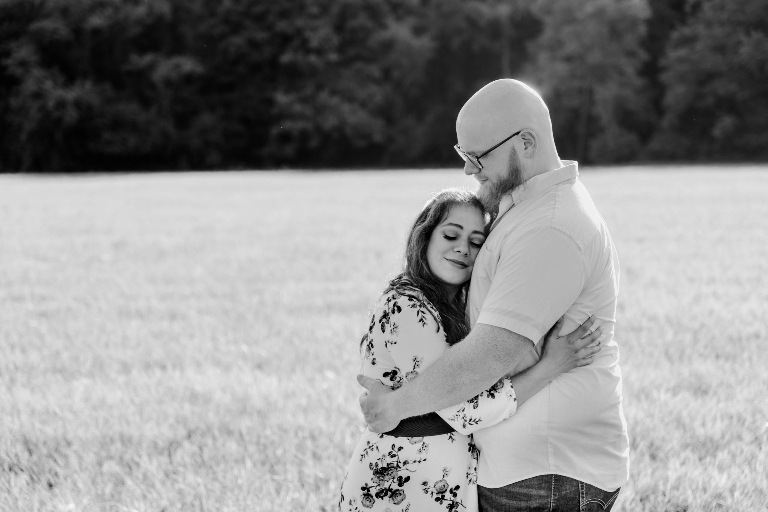Black and white photo of an engaged couple in an open field at East Fork State Park hugging each other with soft smiles.