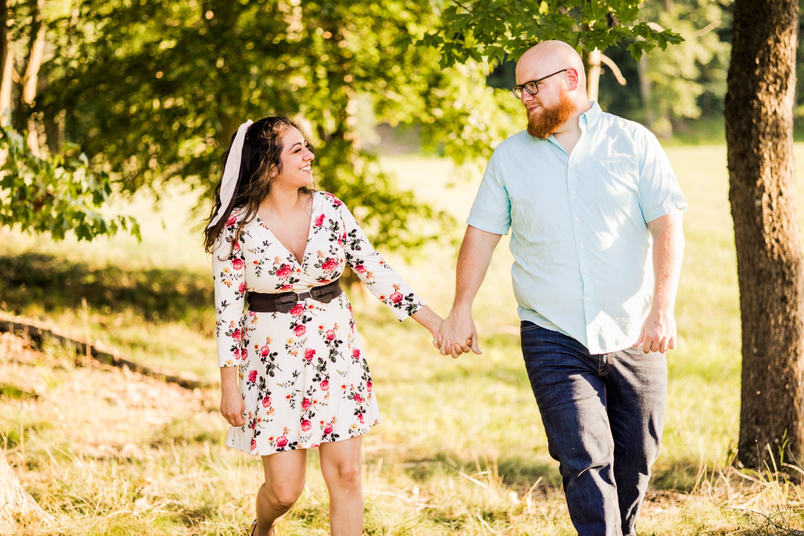 An engaged couple holding hands walking toward the photographer in a grove of tress while smiling and looking at one another in the summer.