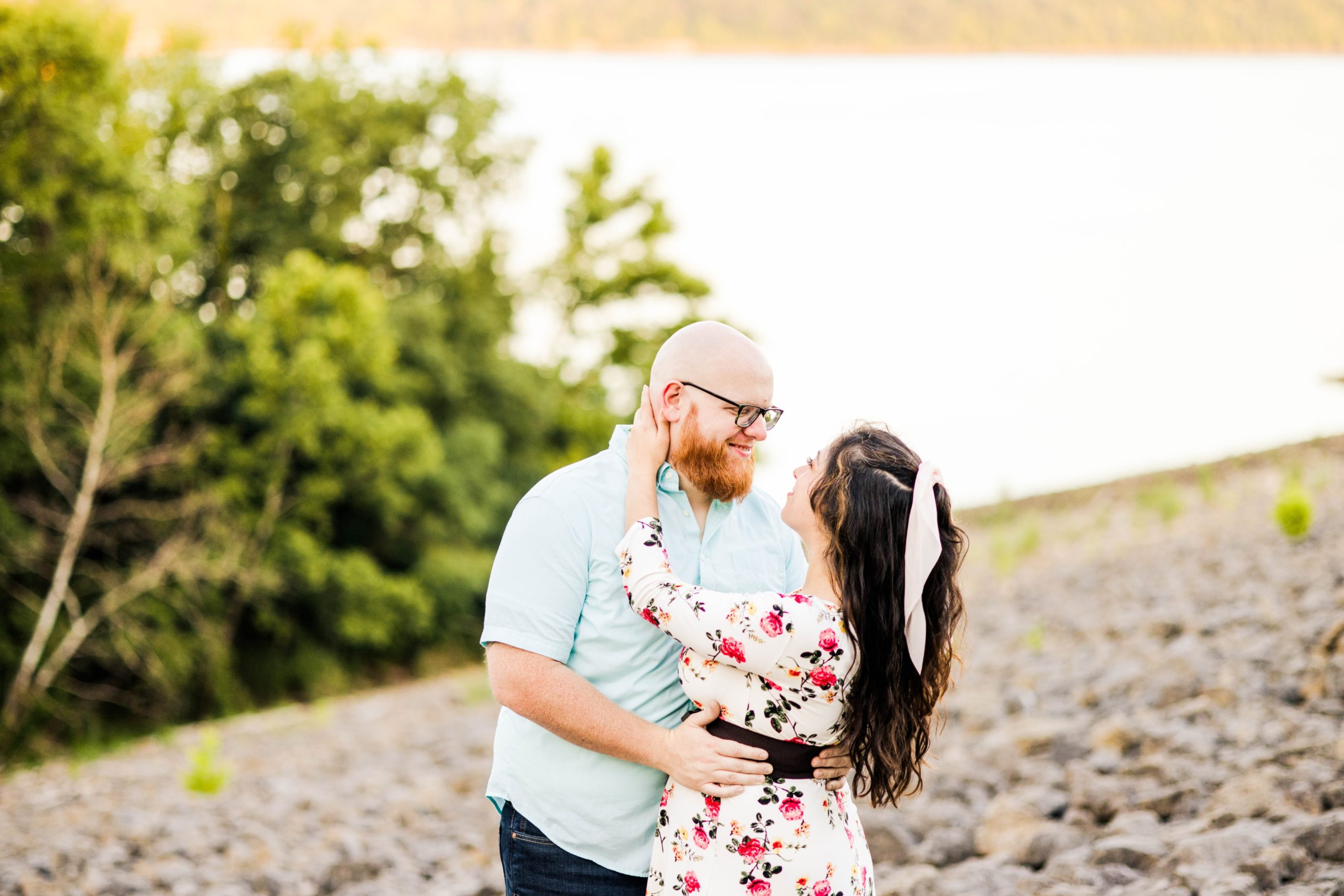Jessica and Sam are looking at each other during their summer engagement photos on the side of a lake in a rock field at sunset time at a local park outside of Cincinnati OH
