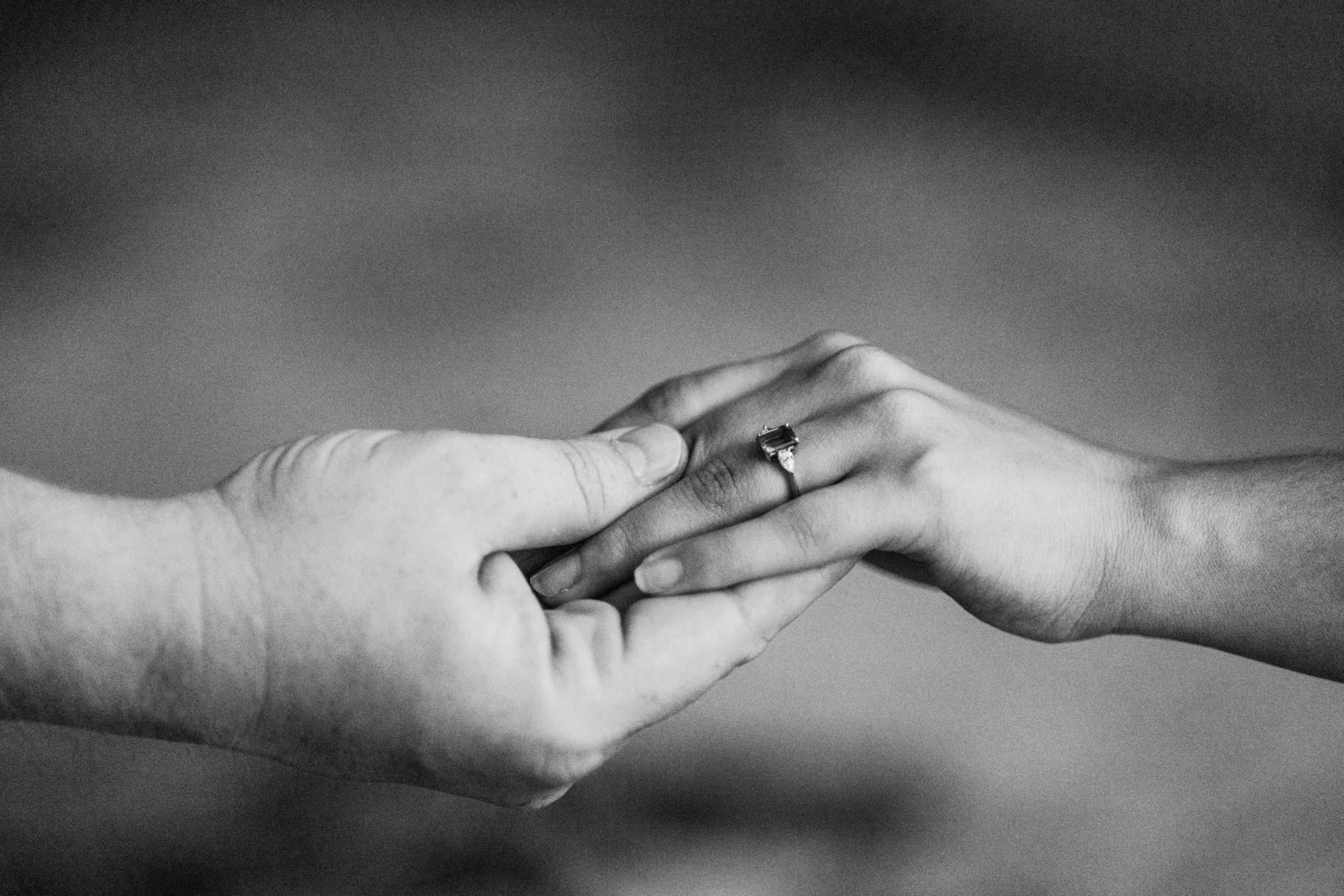 Back and white photo of close up of hands of an engaged couple highlighting the engagement ring with a emerald cut.
