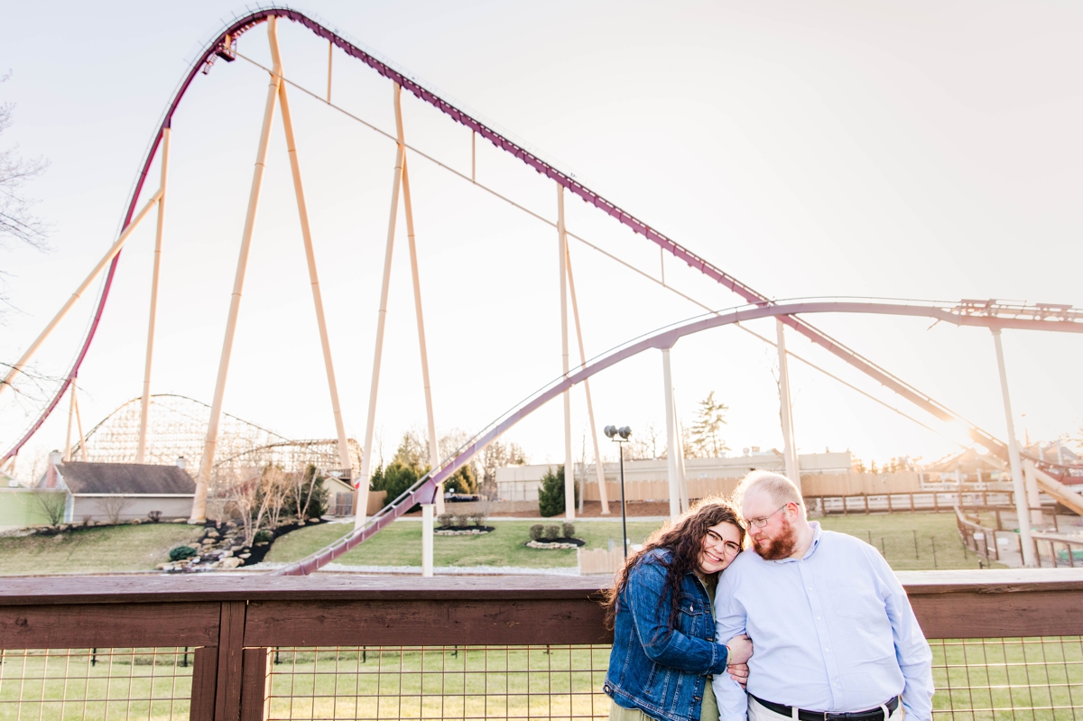 A couple smiling in front of a roller coaster during their engagement session. Follow these tips for engagement photography.
