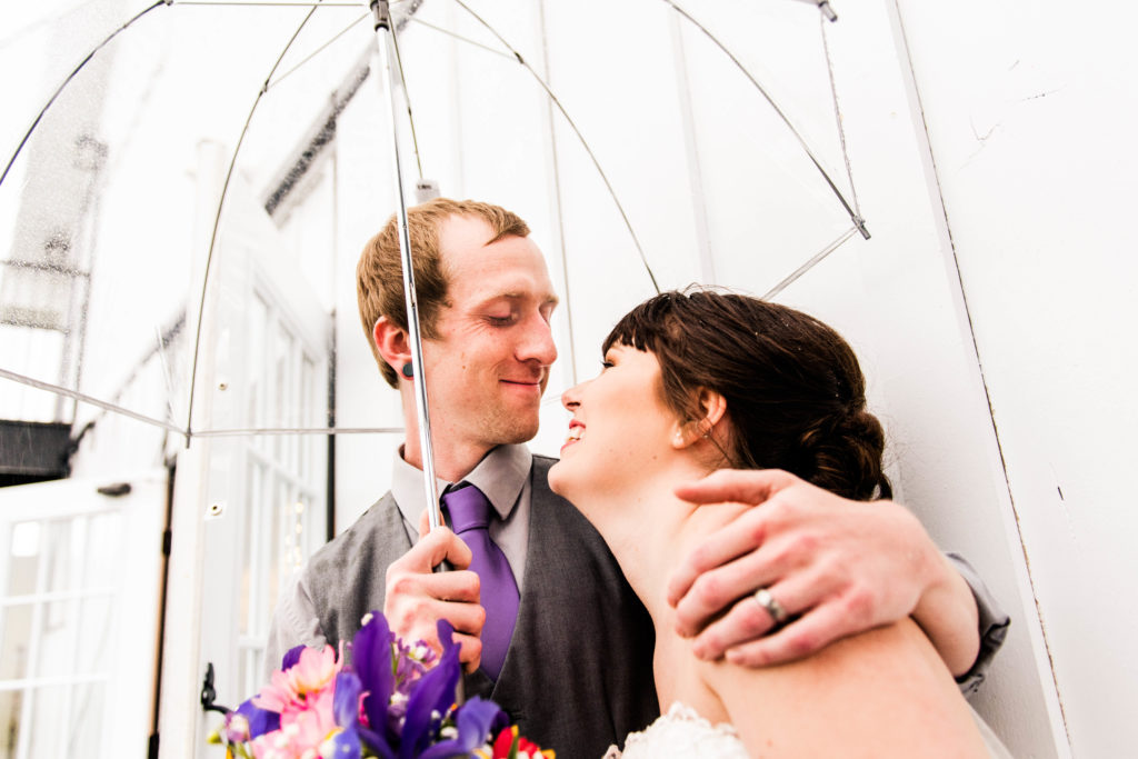 bride and groom leaning against a white building protecting themselves from the rain with a clear umbrella  while smiling at each other on their rainy wedding day