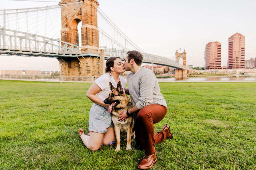 Engaged couple kissing in front of Roebling Bridge with their dog