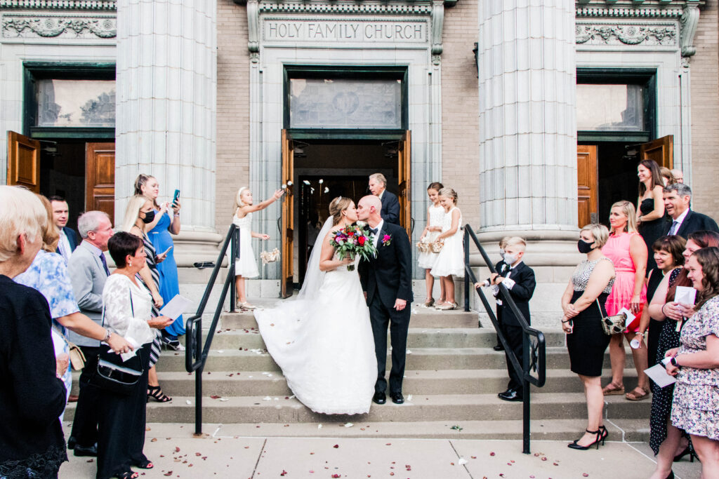 Bride and groom share a kiss on the steps of Holy Family Church, surrounded by wedding guests and flower girls holding baskets of petals, as they prepare for an exciting day navigating multiple wedding locations.