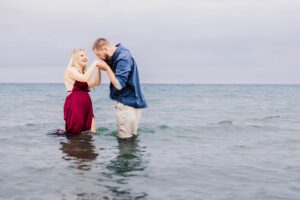 A couple enjoying a sexy beach engagement session, with a man and woman standing in shallow ocean water. The man is kissing the woman's hand while she smiles.