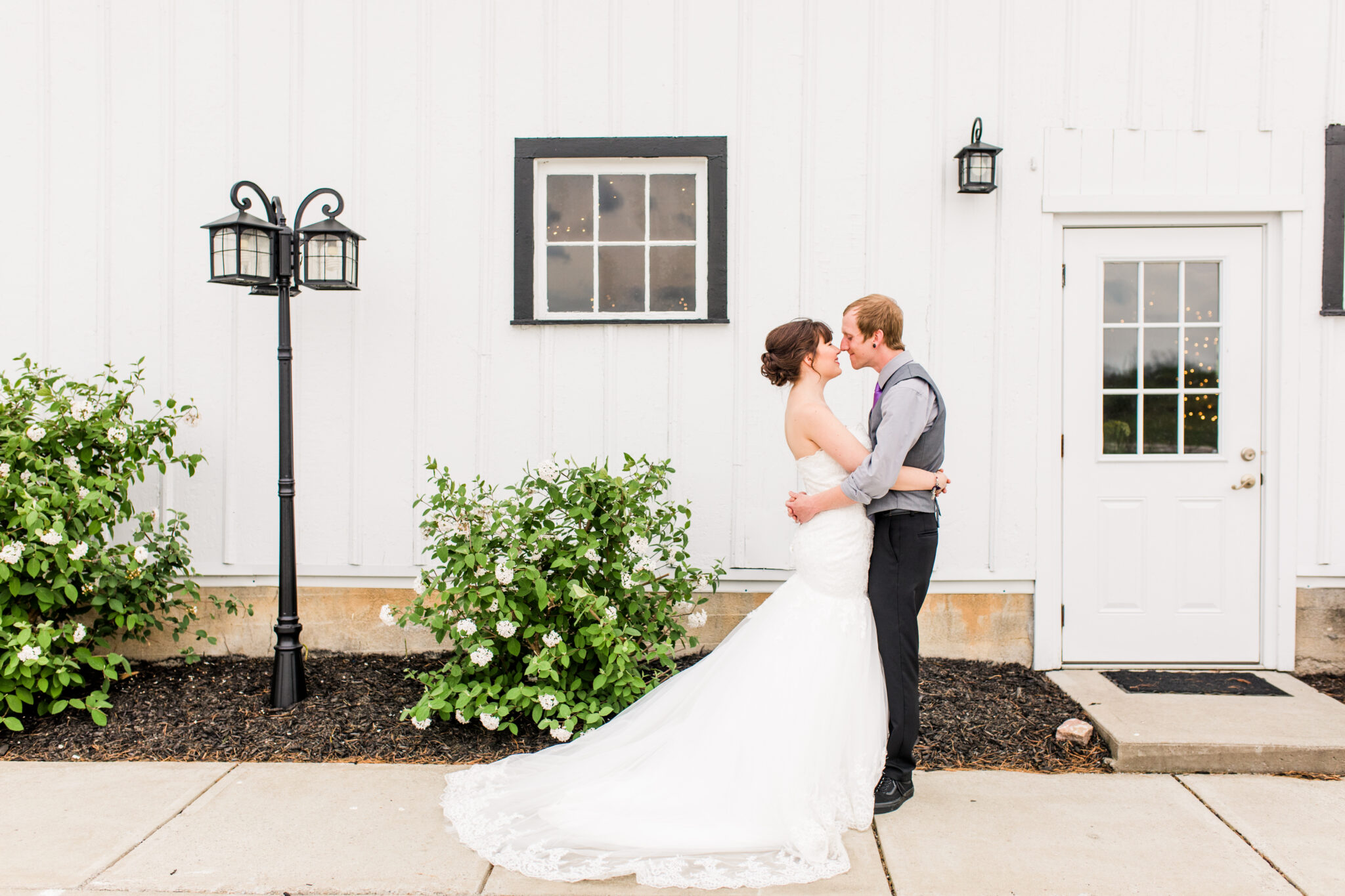A couple in wedding attire shares a kiss in front of the white-paneled building of The Josephina Event Center in Northern Kentucky, complete with a window, door, and two lampposts. Bushes with green leaves are adjacent to them, making for a perfect wedding portrait.