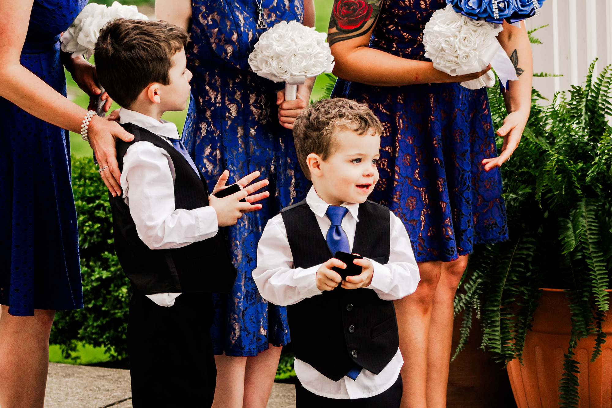 Two boys, children in your wedding party, dressed in formal attire stand in front of women wearing blue lace dresses and holding white bouquets at Pebble Creek Gold Course at a wedding.