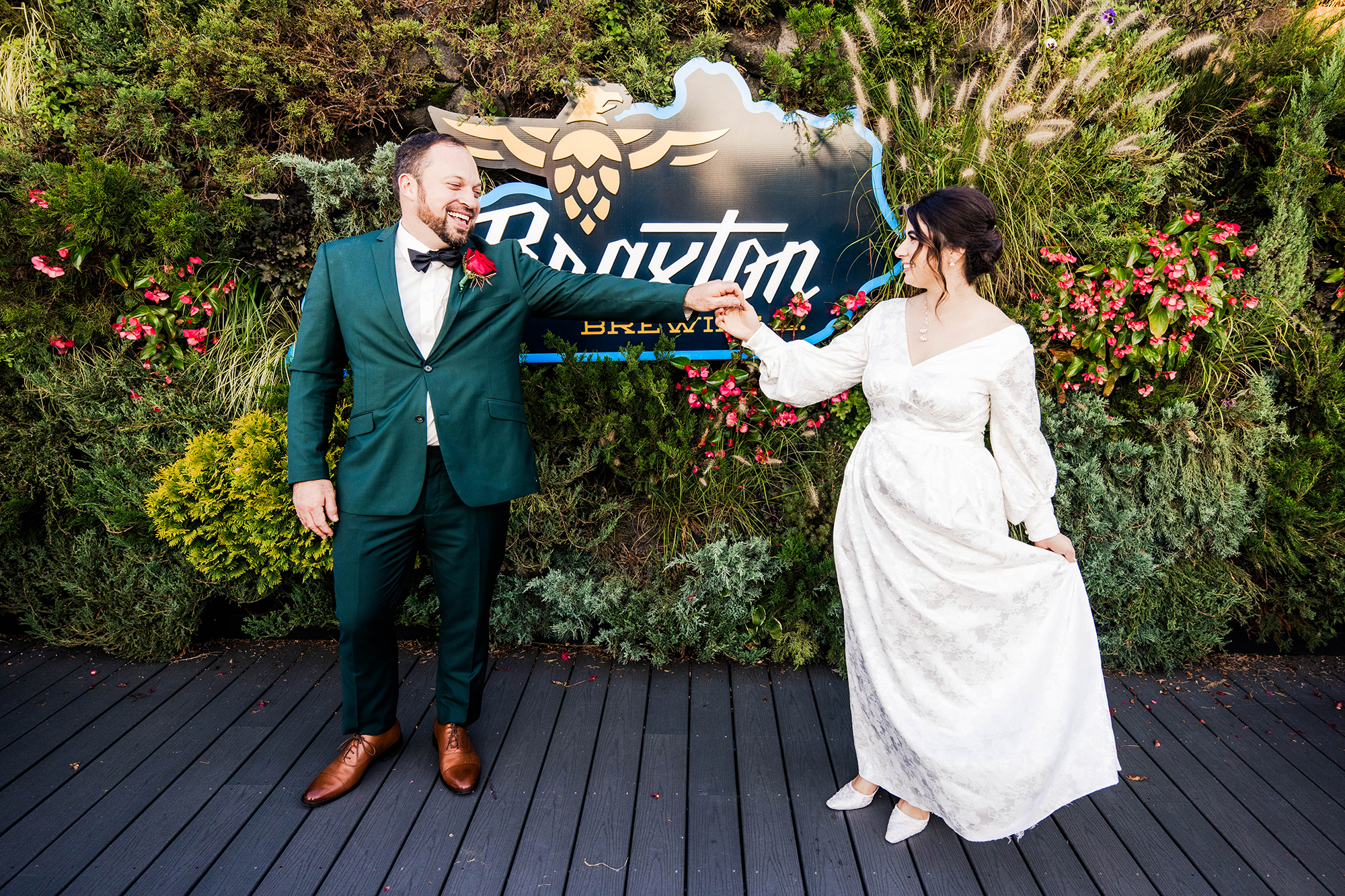 A couple dressed in formal attire poses playfully in front of a brewery sign, surrounded by greenery and flowers, captured beautifully by a Cincinnati wedding photographer.