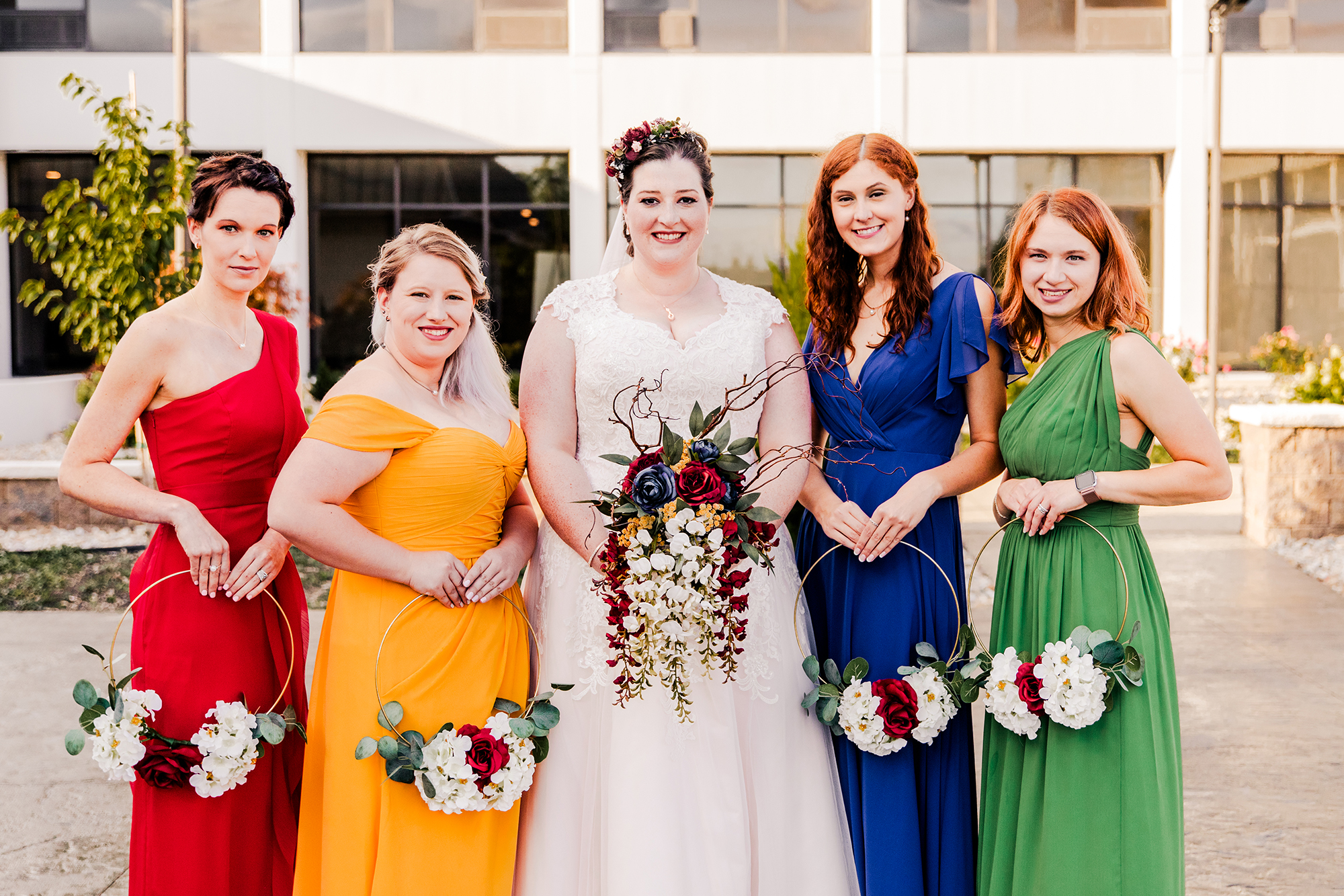 A bride in a white dress stands with four bridesmaids wearing vibrant red, yellow, blue, and green dresses, each holding floral arrangements. This beautiful scene captures the essence of enduring elegance amidst the whispers of dying wedding traditions in an enchanting outdoor setting.