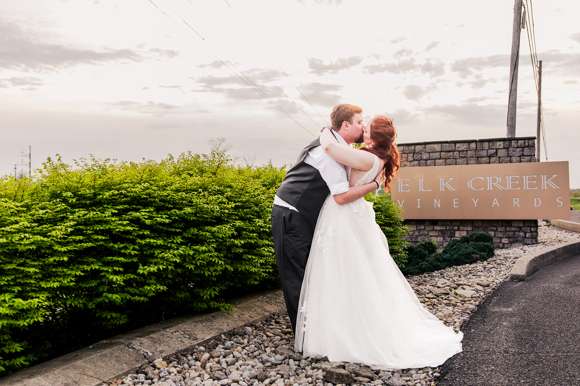 A couple in wedding attire share a tender kiss in front of a sign reading "Elk Creek Vineyards" as the photographer is editing their wedding photos to capture the beauty of their special day.