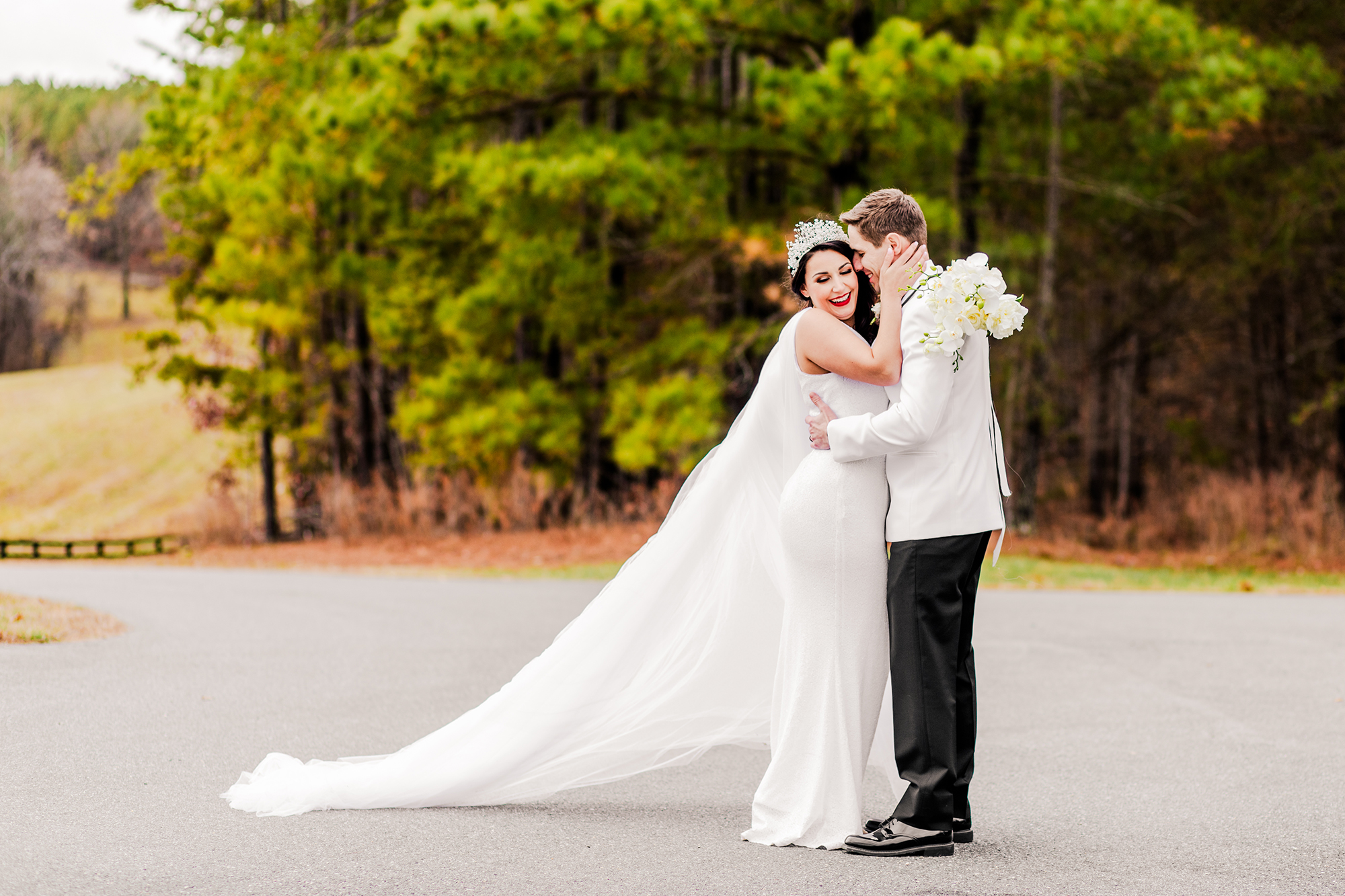 A bride and groom embrace and smile outdoors on a road at Mount Ida Farm, surrounded by trees. The bride wears a long veil and white dress, while the groom holds flowers in his white jacket. They seem eager to get to their reception quickly, savoring this moment of joy before the celebration begins.