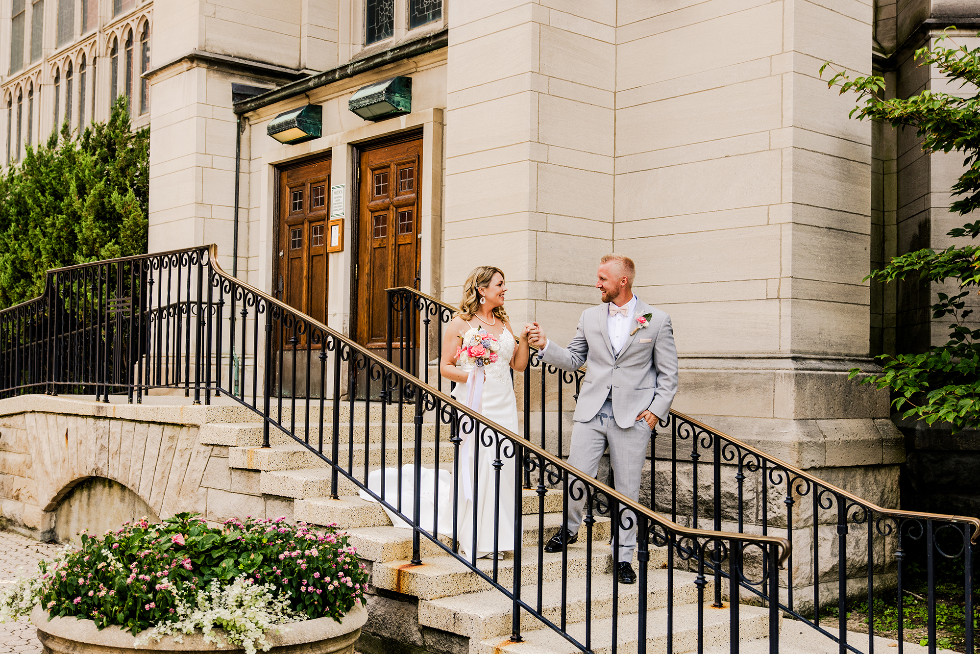 A bride and groom stand on stone steps outside Cathedral Basilica of the Assumption, holding hands and smiling at each other, as they contemplate how many hours they might want their photographer on their wedding day. The groom wears a gray suit, and the bride holds a bouquet in front of the wooden doors.