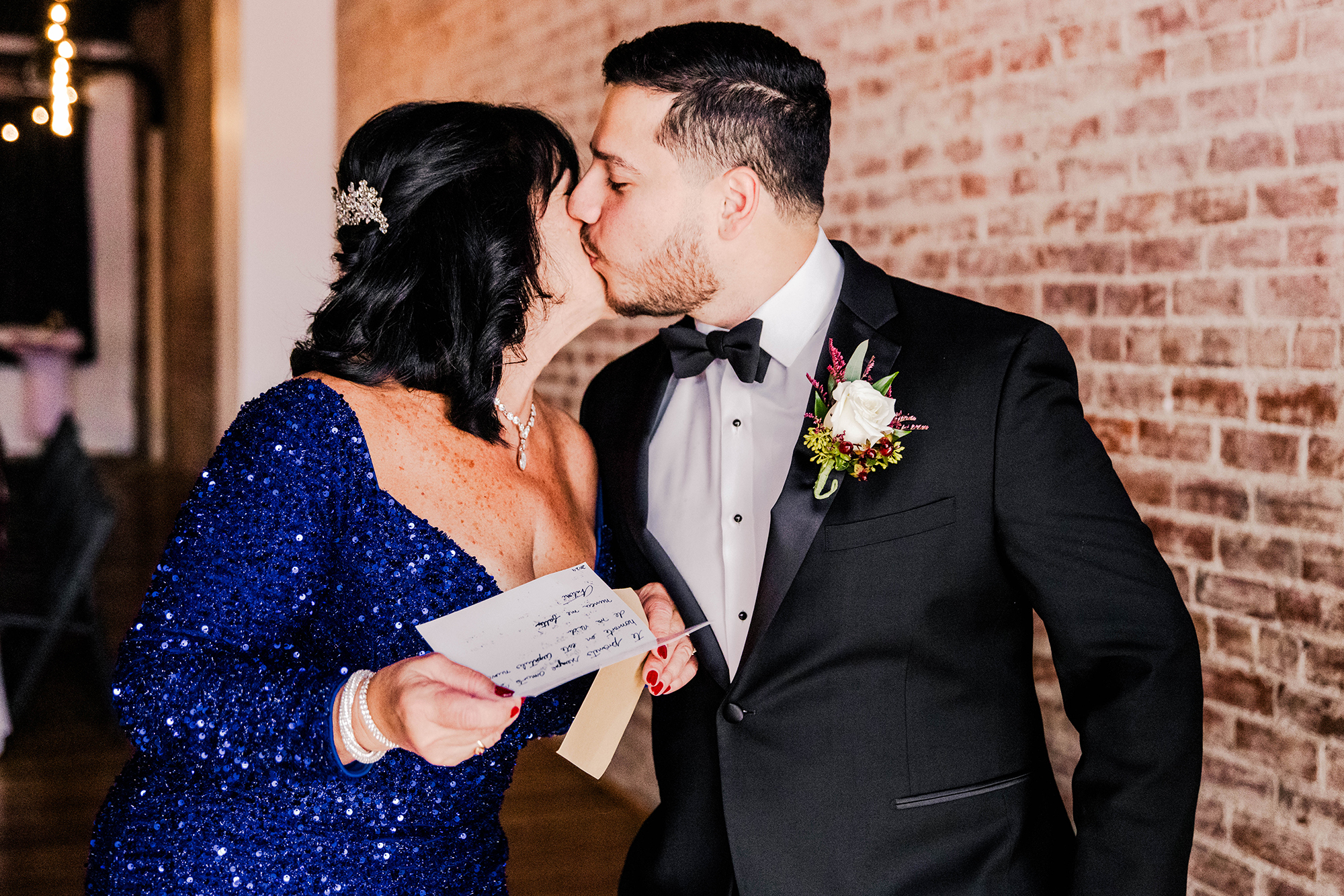 A groom and his mother dressed formally share a kiss in front of a brick wall, capturing one of those cherished mother of the groom moments. The woman, holding a heartfelt card from her son, smiles warmly at this tender memory in the making.