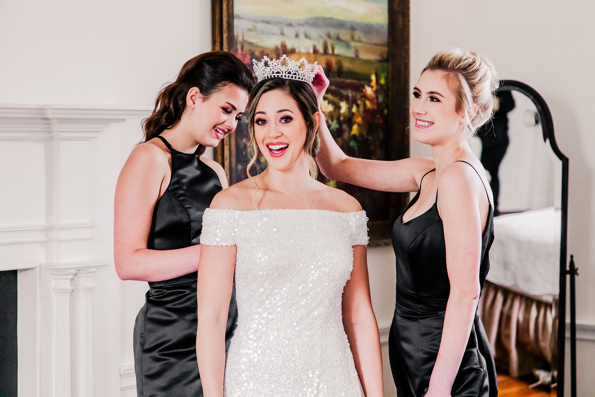 Two women in black bridesmaids dresses adjust a tiara on a smiling bride in a white gown at Mount Ida Farm, who is respecting her wedding party, inside a room adorned with a painting and mirror.