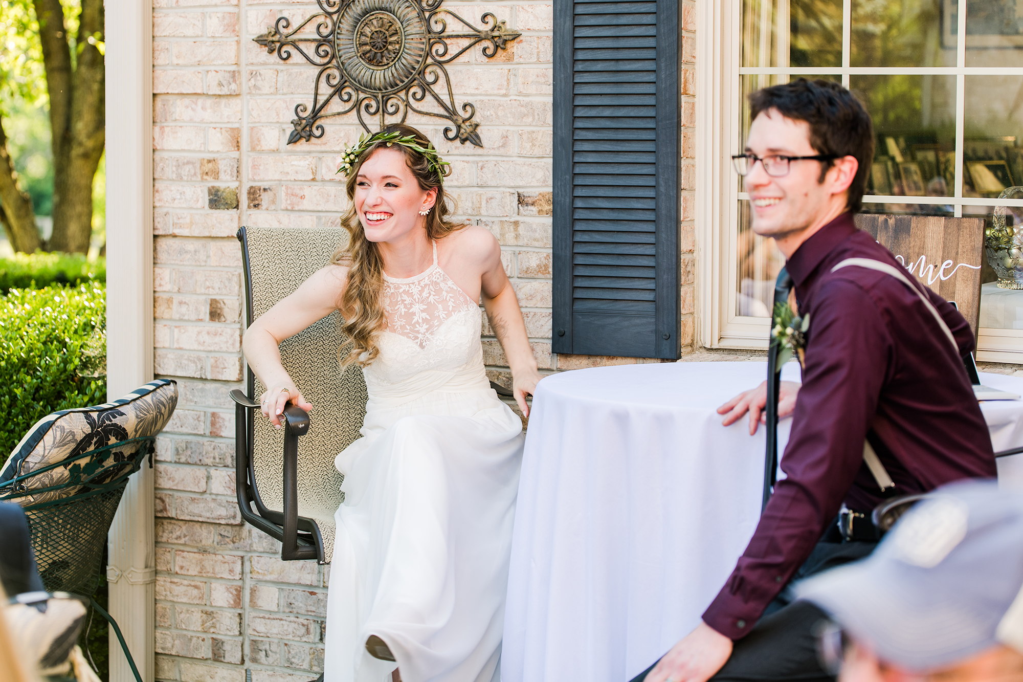 At a small, intimate wedding, a woman in a wedding dress and a man in glasses, dressed in a dark shirt, sit outdoors at a table adorned with white cloth against a brick wall—capturing the essence of candid wedding photography.