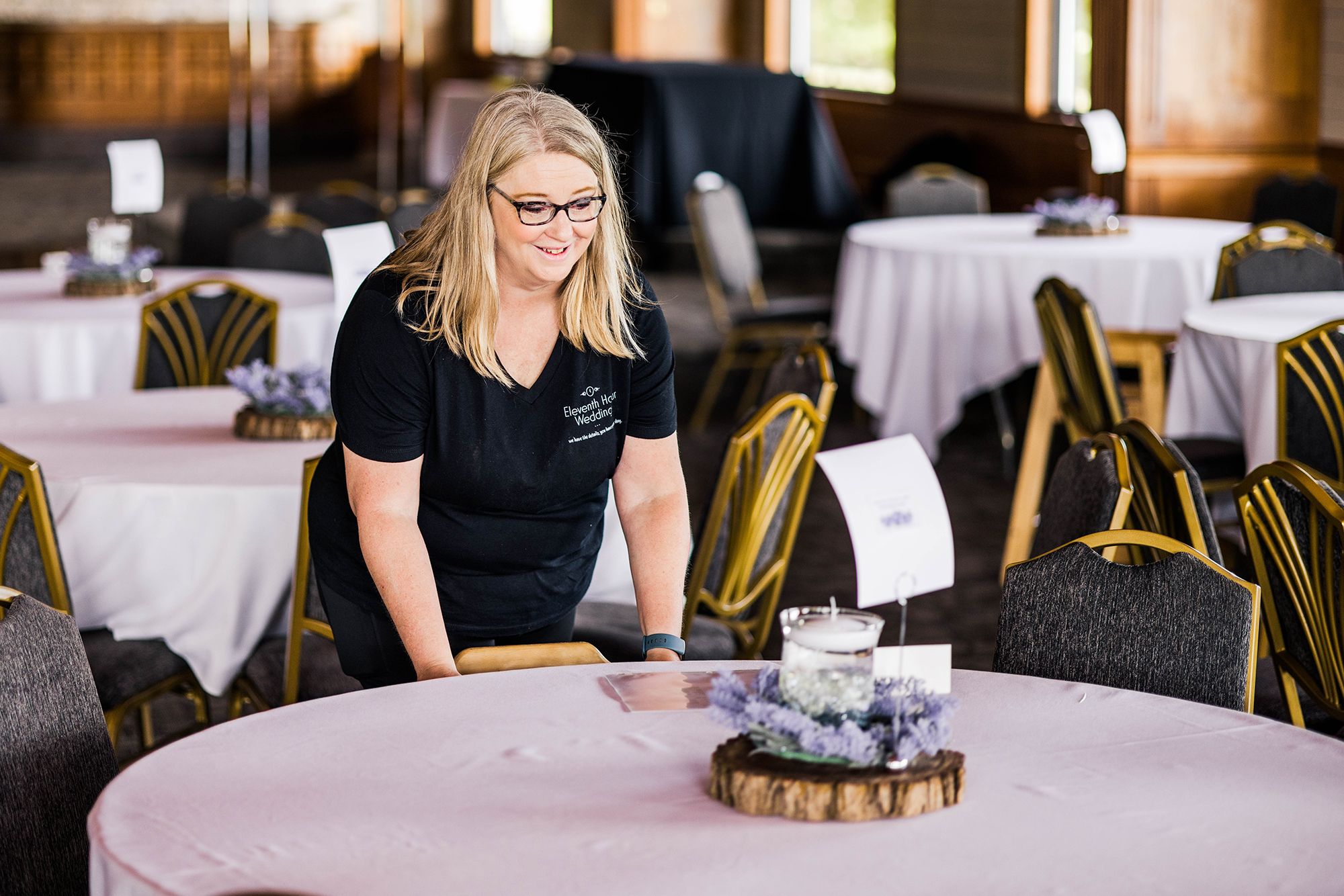 A wedding planner with 11th Hour Weddings and Events elegantly sets a round table with a purple tablecloth and a decorative centerpiece, transforming the room's empty chairs into an inviting celebration space. Perfect example of a wedding planner or a wedding coordinator.