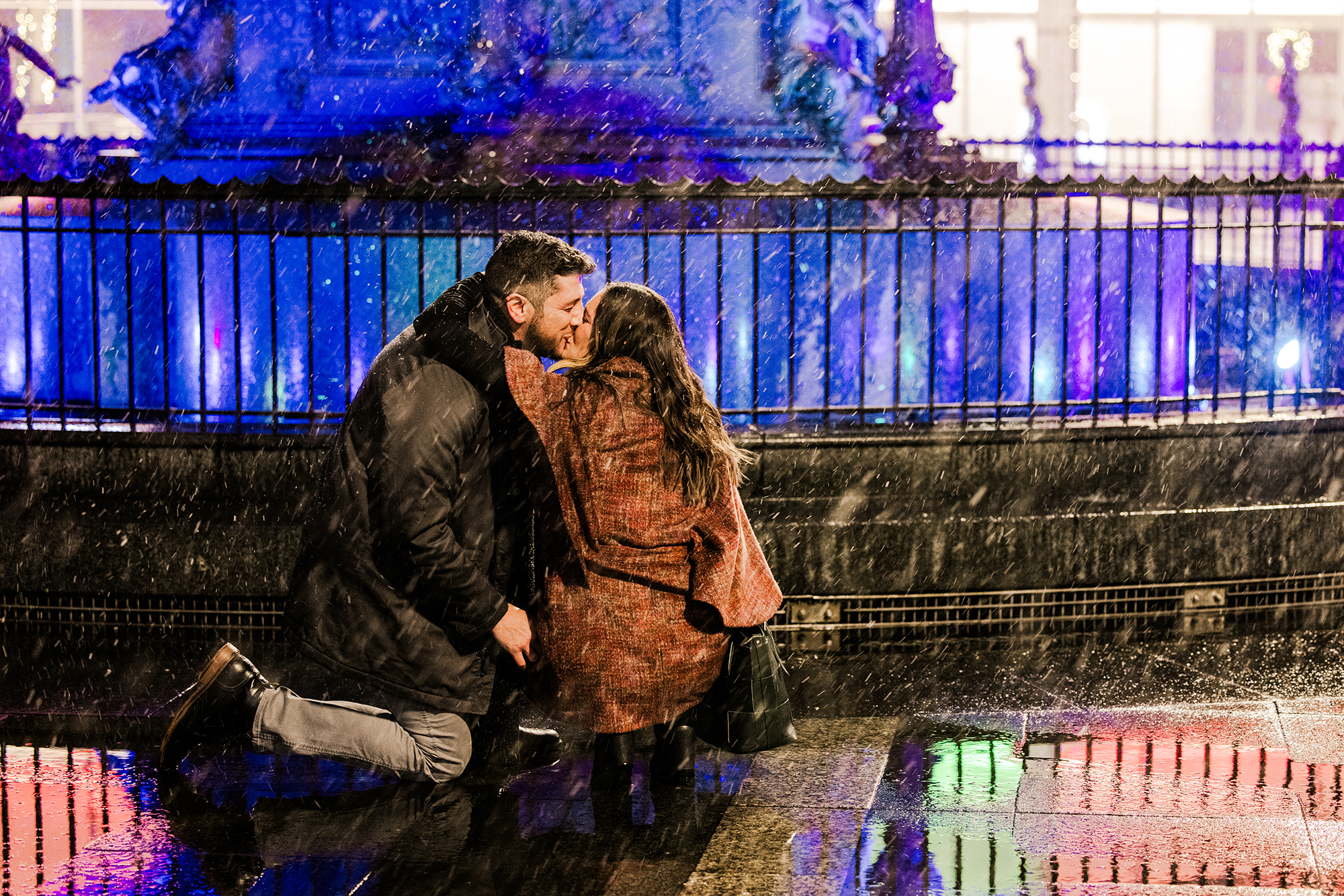 You're Enageged. what's next? A couple, newly engaged and dreaming of what’s next, shares a kiss in the rain, illuminated by colorful lights in the background.