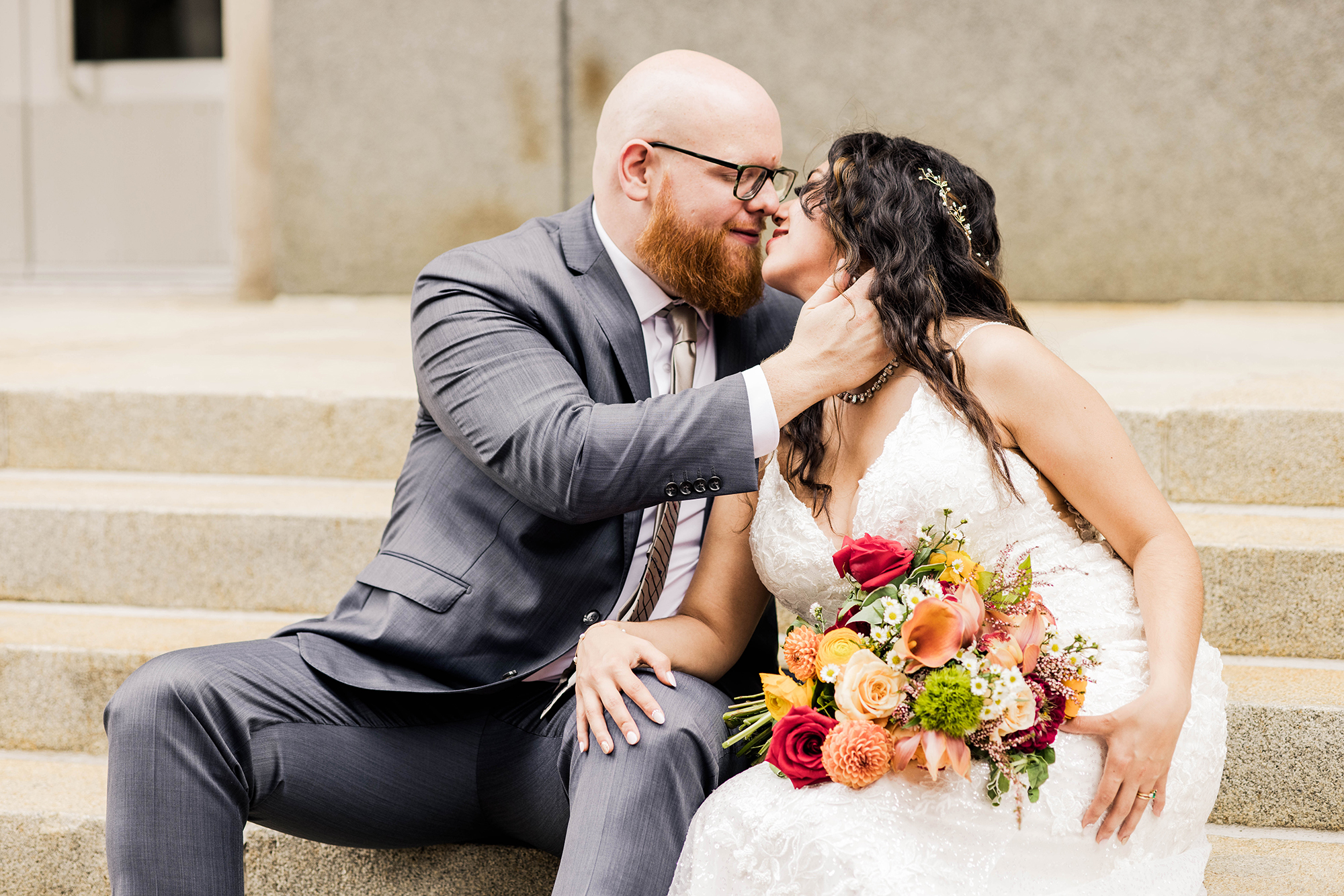 A couple dressed in wedding attire shares a tender moment on stone steps, captured effortlessly by a Cincinnati wedding photographer. The bride holds a bouquet of colorful flowers, ensuring they look natural and not be awkward in photos.