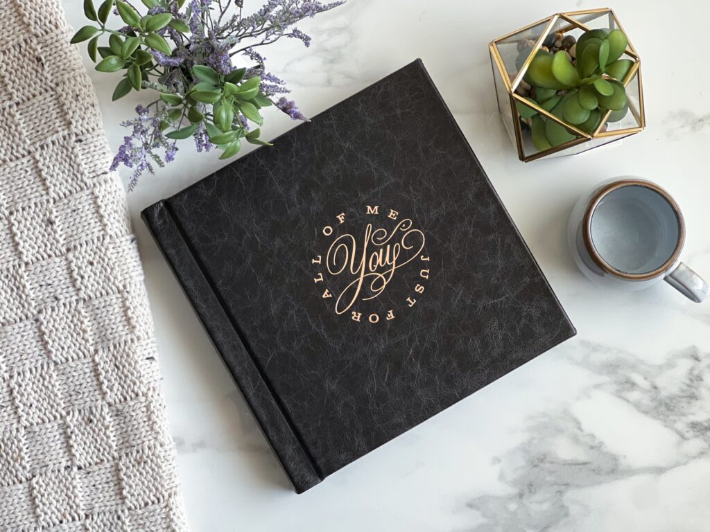 A black boudoir album with gold lettering that says "All of Me Just for You" sits on a marble table next to a knitted cloth, green plants, and a gray mug—an elegant Cincinnati wedding gift idea.